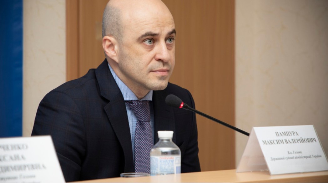 На фото чоловік без волосся у темному костюмі та краватці, сидить за столом з мікрофоном. Біля нього пляшка води паперова табличка з ім’ям Пампура Максим Валерійович dqxikeidqxitkant